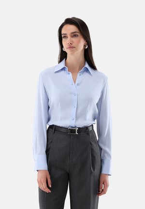 Femme aux longs cheveux bruns portant une chemise bleu clair boutonnée rentrée dans un pantalon taille haute gris foncé avec une ceinture noire, debout sur fond blanc.