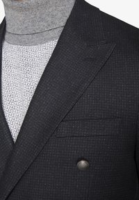 Grå turtleneck under en mørk mønstret blazer. Blazeren har et dobbeltradet design og en stiklomme med metalknapdetalje.