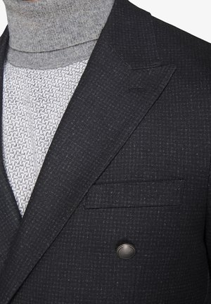 Suéter gris de cuello alto debajo de un blazer oscuro con un patrón. El blazer tiene un diseño de doble botonadura y un bolsillo de parche con detalle de botón metálico.