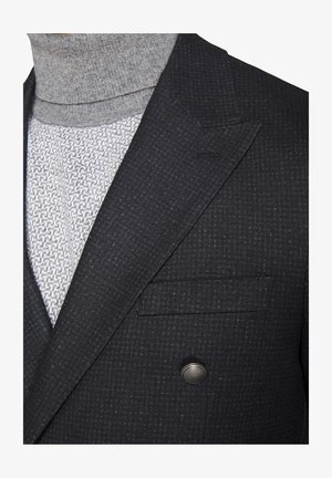 Gray turtleneck beneath a dark patterned blazer. Blazer features a double-breasted design and a patch pocket with metallic button detail.