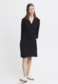 Schwarzes, knielanges Hemdkleid mit langen Ärmeln, V-Ausschnitt und gebundenem Taillenbereich. Aus glattem Stoff, getragen mit weißen Sneakers.