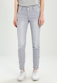 Pantalones ajustados en blanco con un patrón de diamantes azules. Cuentan con bolsillos frontales y cremalleras en los tobillos. Combinados con zapatillas blancas.