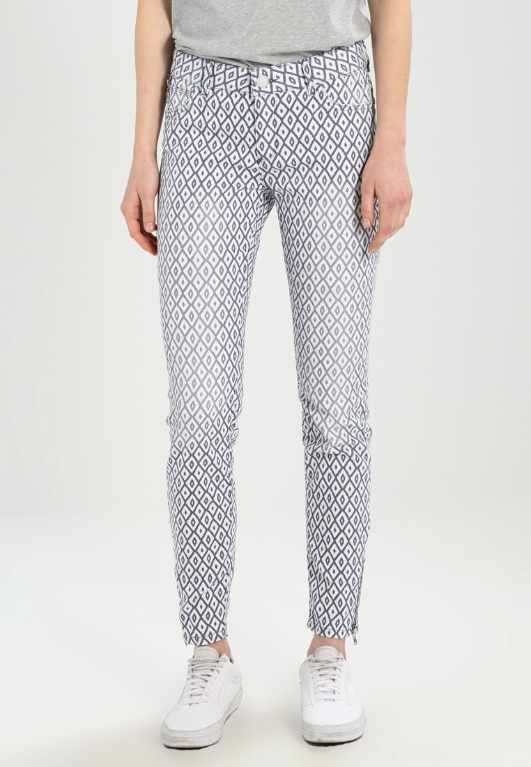 Pantalones ajustados en blanco con un patrón de diamantes azules. Cuentan con bolsillos frontales y cremalleras en los tobillos. Combinados con zapatillas blancas.