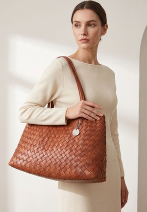 Femme en robe crème tenant un grand sac cabas en cuir tressé marron avec des anses et une étiquette métallique circulaire.