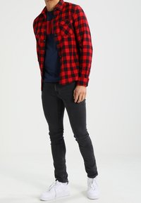 Camisa de franela de cuadros rojos y negros, de manga larga, con dos bolsillos en el pecho, puesta sobre una camiseta azul marino. Combinada con jeans negros ajustados y zapatillas blancas.