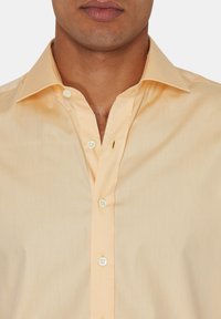 Ljus persikofärgad skjorta med krage. Har en slät tygtextur och fem vita knappar längs framden.