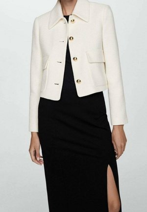 Femme portant une veste courte blanche à boutons dorés et une robe noire avec une fente haute sur la cuisse.