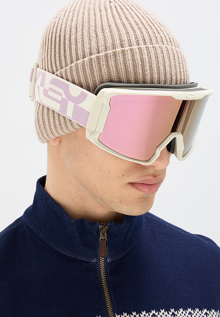 Skibrille mit einem weißen Rahmen und pinkem verspiegeltem Glas, ausgestattet mit einem gemusterten elastischen Riemen. Getragen mit einer beigefarbenen Strickmütze und einem dunklen Reißverschlusshemd.