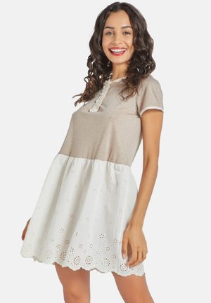 Mujer sonriente con cabello rizado que lleva un vestido beige y blanco, con borde de encaje y dobladillo de ojales, posando con confianza con una mano en la cadera.