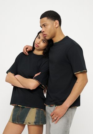 Jeune homme et femme portant des chemises noires avec une bordure camouflage, la femme s'appuyant sur l'épaule de l'homme, debout devant un fond uni.