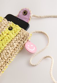 Prepletena slamnikova torbica čez ramo z rumenimi črtami in roza črtasto preklopno zaponko, ki drži črni pametni telefon, roza obesek nasmejanega obraza na bež vrvici za nošenje.