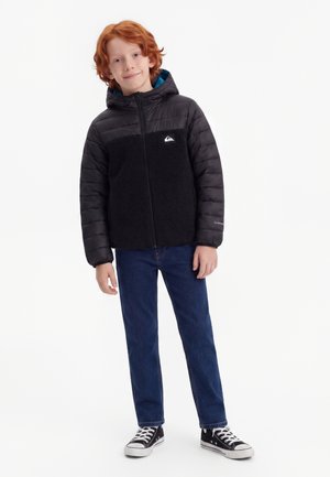 Jeune garçon roux portant une veste à capuche noire, un jean bleu et des baskets noires, debout devant un fond blanc.