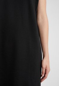 Musta hihaton mekko, jossa on sileä ja pehmeä rakenne. Ominaisuuksia ovat rento leikkaus ja polvitason helma, jossa käsi on näkyvissä helman lähellä.