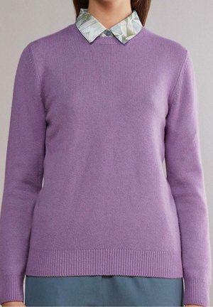 Vrouw die een lichtpaarse gebreide trui draagt over een overhemd met een bloemenpatroon, gecombineerd met een blauwe broek, alleen de romp zichtbaar.