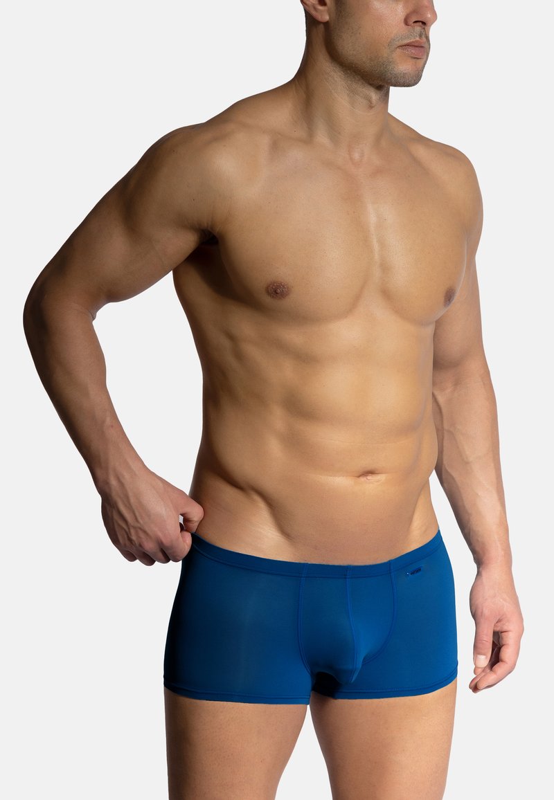 Blue boxer briefs made of smooth fabric, featuring a snug fit, flat waistband, and contrasting stitching detail. Model stands with one hand adjusting.