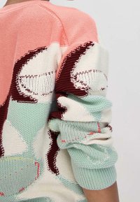 Pull en tricot avec un motif multicolore comprenant du rose pastel, du menthe et du bordeaux, des fils texturés, et une manche confortable retroussée.