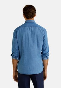 Camisa vaquera en azul claro, mangas largas remangadas, cuello clásico, textura suave, puños con un solo botón y ajuste entallado en la parte posterior.