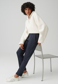 Pullover bianco in pile con zip a metà e maniche larghe, abbinato a pantaloni scuri a trama e sneakers bianche con accenti grigi.