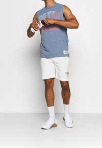 Camisa sin mangas azul con el texto "HACE EL TRABAJO", combinada con shorts blancos que tienen un logo marrón claro. Zapatillas blancas y botella de agua en la mano.