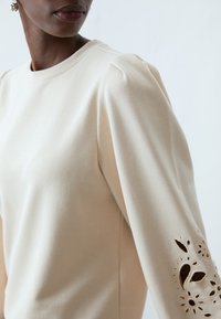 Maglietta beige a maniche lunghe con scollatura rotonda, caratterizzata da dettagli floreali intagliati sulle maniche e una texture liscia e morbida.