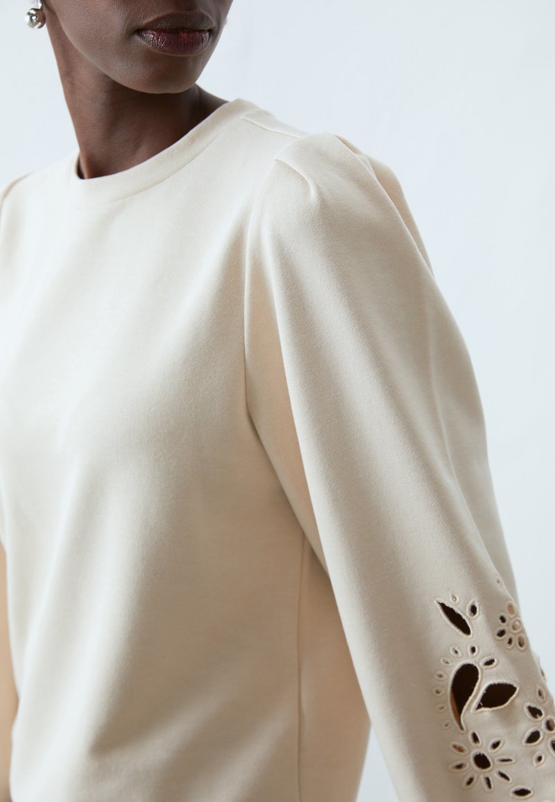 Maglietta beige a maniche lunghe con scollatura rotonda, caratterizzata da dettagli floreali intagliati sulle maniche e una texture liscia e morbida.