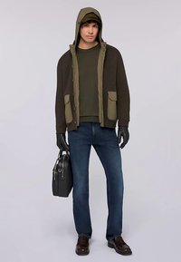 Chaqueta verde oliva con textura acanalada, dos bolsillos delanteros, superpuesta sobre una camisa verde oscuro. Vaqueros oscuros, guantes negros y un bolso negro.