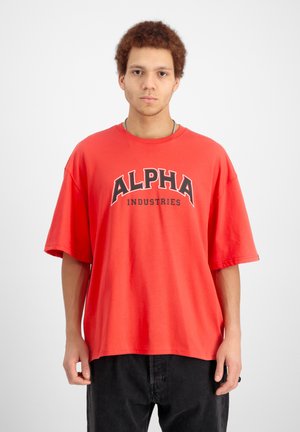 Young man with curly hair wearing a loose red T-shirt with "Alpha Industries" text and black pants, standing against a plain white background.