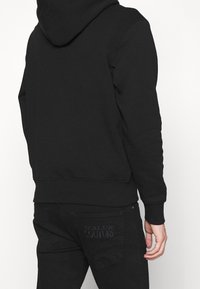 Osoba v černé mikině s kapucí a černých džínách, s výšivkou "Versace Jeans Couture" na zadní kapse, zobrazená zezadu.