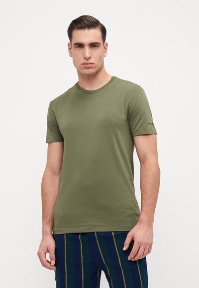 Olive green t-shirt in cotton fabric, short sleeves, round neckline, featuring subtle branding on the sleeve. Model paired with striped pants.