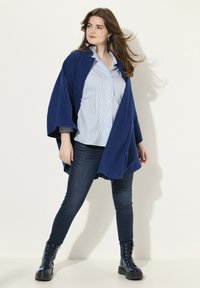 Cardigan sovradimensionato di un blu scuro con maniche ampie, indossato sopra una camicia a righe blu chiaro. Abbinato a jeans scuri e stivali stringati lucidi.