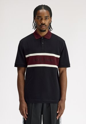 Homme aux cheveux tressés portant un polo noir avec une bande horizontale bordeaux et blanche et le logo "Fred Perry", debout devant un fond uni.