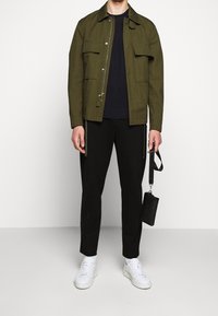 Bawełniana kurtka w oliwkowej zieleni z dwoma przednimi kieszeniami, w połączeniu z granatową koszulą, czarnymi spodniami i białymi trampkami, trzymająca czarną saszetkę.