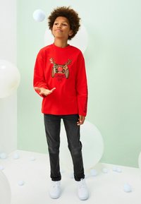 Red long-sleeve shirt with a game controller graphic featuring antlers, paired with black pants and white sneakers. Set against a pastel backdrop.