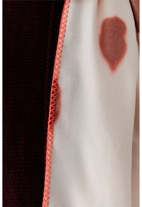 Deux morceaux de tissu reliés par une couture orange vif ; l'un en tricot de couleur marron foncé et l'autre lisse blanc avec des taches brun-rouge.