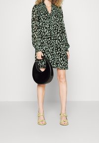 Vihreä ja musta kuvioitu mekko, jossa on röyhelökaulus, pitkät hihat ja nappikiinnitys. Pitelee mustaa käsilaukkua ja käyttää vaaleita sandaaleja.