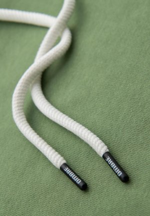 Lazos trenzados blancos con puntas negras que presentan el texto "DUMBARKE," reposando sobre un fondo verde texturizado.