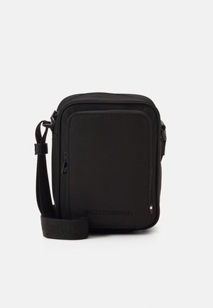 Zwarte rechthoekige crossbodytas met voorvak met ritssluiting, verstelbare band en klein Tommy Hilfiger-logo nabij de rechteronderhoek.