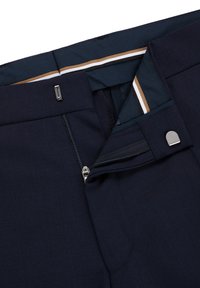 Marineblaue Hose mit einem Reißverschluss und silbernem Zubehör. Sie verfügt über eine glatte Textur und ein kontrastierendes, gestreiftes Innendetail des Bunds.