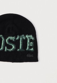 Musta neulotusta materiaalista valmistettu pipo, jossa on hulppeat mintunvihreät kirjainmerkit muodostaen tekstin "LACOSTE" ja pieni musta logokoriste reunuksessa.