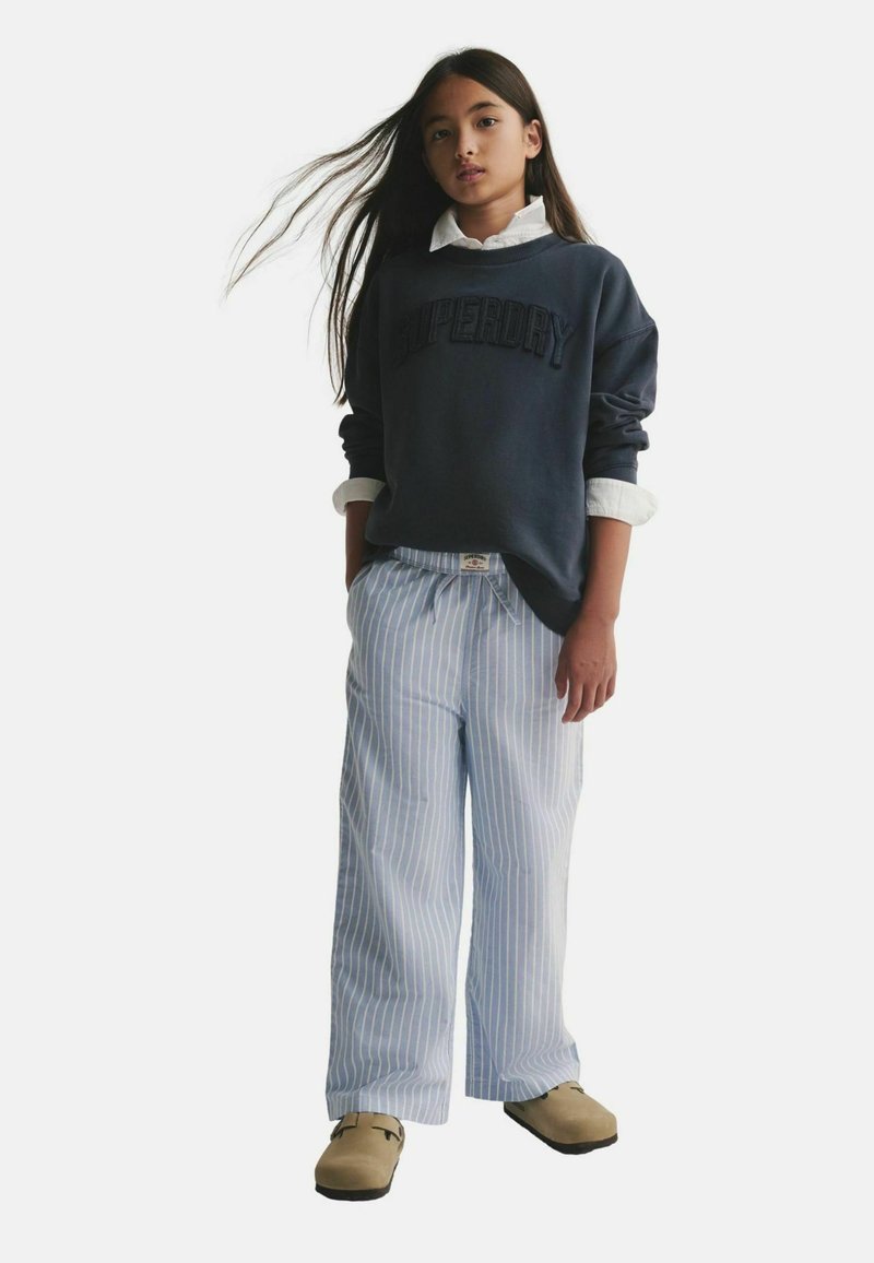 Dunkelblaues Sweatshirt mit "Superdry"-Schriftzug, weiße Bluse darunter, gestreifte hellblaue weit geschnittene Hose und beigefarbene Schuhe.