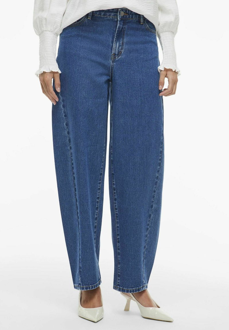 Platas kāju zilas denima bikses ar augstu jostasvietu, apdarināta apmale un klasiskā piecu kabatu dizains, saskaņotas ar bēšiem augstpapēžu apaviem.