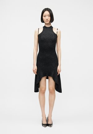 Kvinde stående oprejst iført en ærmeløs, sort kjole med høj hals og asymmetrisk kant samt sorte spidse hæle på hvid baggrund.