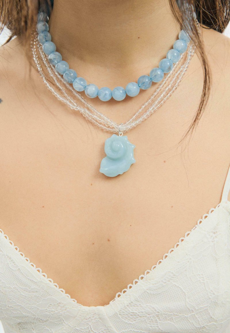 Deux colliers : l’un avec de grosses perles rondes bleu clair, l’autre avec des perles transparentes et un pendentif coquillage bleu clair sur une femme portant un haut en dentelle blanche.