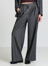 Pantalon gris à jambes larges avec plis, fabriqué en tissu texturé. Caractérisé par une taille haute et des poches avant, associé à des chaussures noires brillantes.