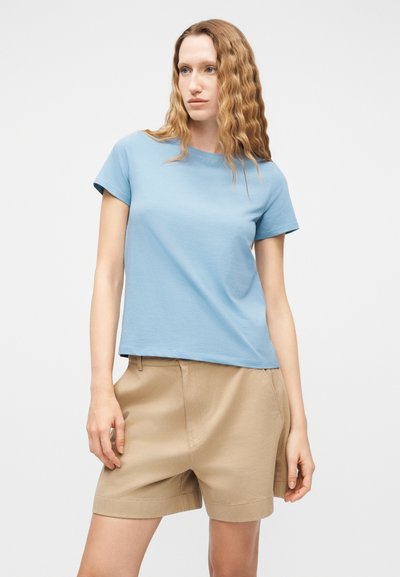 Γυναίκα με κυματιστά ξανθά μαλλιά, φορώντας ανοιχτό μπλε κοντομάνικη μπλούζα και μπεζ σορτς μέχρι το γόνατο, στέκεται μπροστά σε απλό λευκό φόντο.