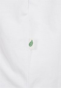 Material alb cu o mică etichetă de culoare alb-crem, având o ilustrație cu o frunză verde, cusută de-a lungul cusăturii centrale.