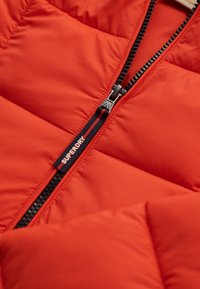 Doudoune rouge à design matelassé, avec une fermeture éclair noire et une étiquette logo "Superdry" près de la fermeture. Texture lisse, matériau léger.
