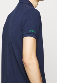 Granatowa koszulka polo o dopasowanym kroju. Posiada prążkowany kołnierzyk, krótkie rękawy oraz zielone logo "Polo" wyszyte na lewym rękawie.