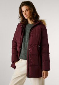 Chaqueta acolchada burdeos con cremallera frontal, cierres a presión y capucha de pelaje sintético. Presenta paneles acolchados y bolsillos laterales. Llevada sobre un suéter gris.