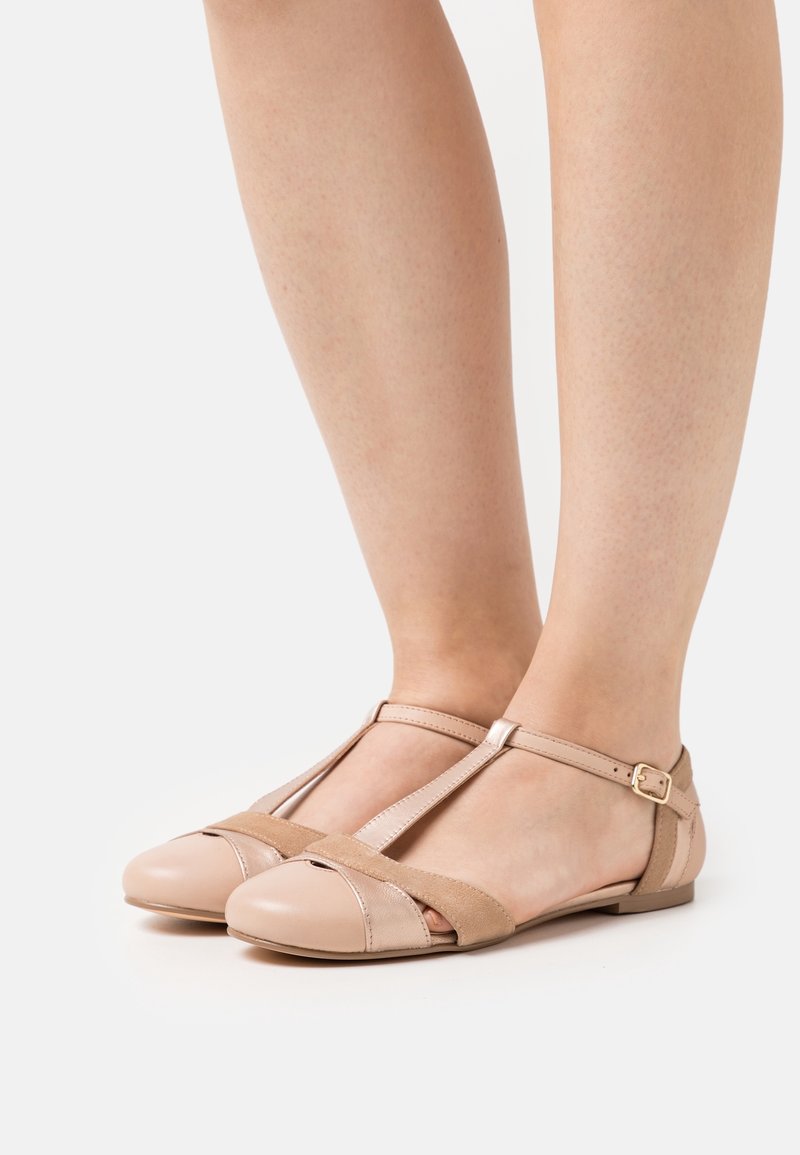 Sandales plates beiges à bride en T avec une petite boucle, portées pieds nus, laissant apparaître la peau à travers l'orteil ouvert et les découpes latérales.